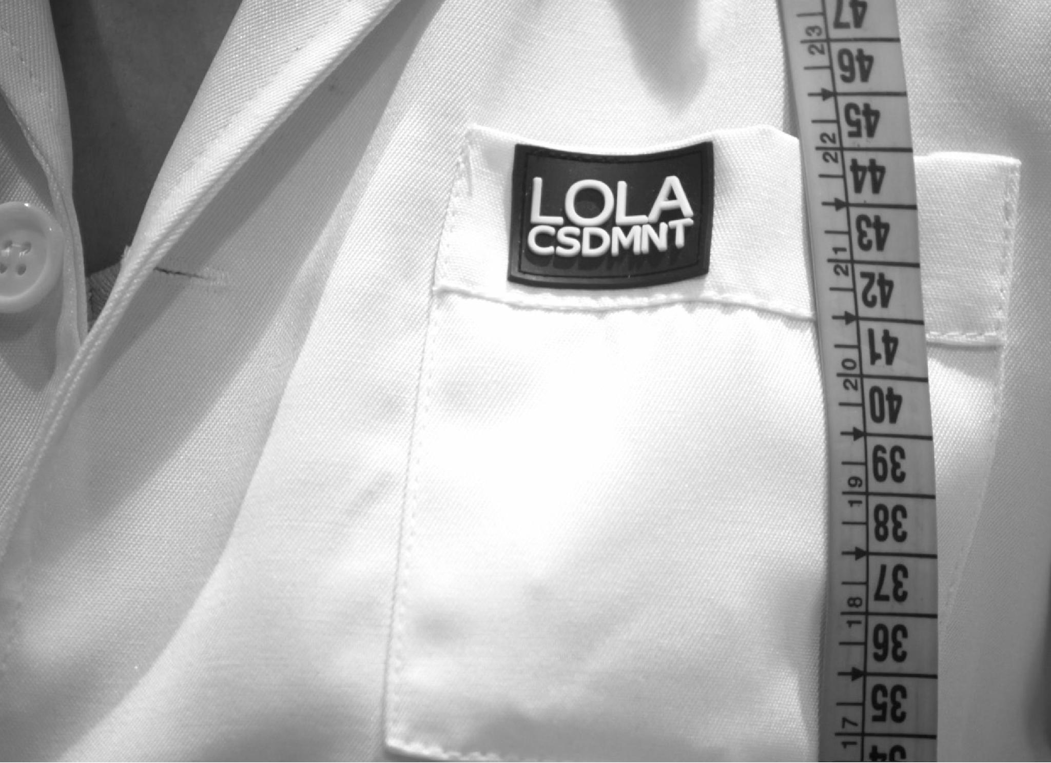 Detalle de un bolsillo de camisa de tela blanca, en el que hay una etiqueta con el texto 'Lola CSDMNT'. Colgando en vertical, pasando sobre el bolsillo hay una cinta de sastre.